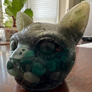 以下が含まれることがあります： 緑と黒の猫の彫刻。樹脂製で、緑と白の石が埋め込まれている。彫刻はリアルな猫の顔で、鼻と目が細かく作られている