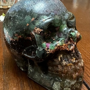 May include: A black skull candle with green and pink crystals embedded in the wax. The skull is decorated with gold leaf accents.