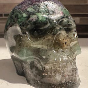 May include: A green, purple, and black skull made of crystal. The skull is translucent and has a shiny finish.