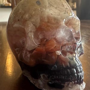 May include: A clear quartz crystal skull with a dark purple and red interior. The skull is carved in detail and has a smooth, polished finish.