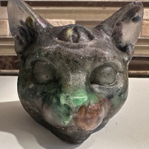 May include: A black, green, and brown resin cat sculpture with a third eye on its forehead. The cat's eyes are closed and its mouth is slightly open.