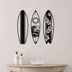 May include: Three black metal surfboard wall decor pieces with different designs. The center surfboard has a sun, waves, and a cloud design. The surfboard on the left is plain. The surfboard on the right has a floral design.