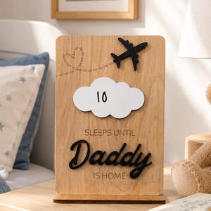 May include: A rectangular wooden countdown sign featuring a black airplane silhouette, a dashed heart flight path, and a white cloud cutout with the number 10. The text reads SLEEPS UNTIL Daddy IS HOME in black lettering on the light wood surface.