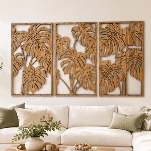 May include: A set of three rectangular wooden wall panels featuring intricate laser-cut monstera leaf designs. Each panel is framed in wood and displays a natural brown grain, arranged horizontally to create a cohesive botanical art display.