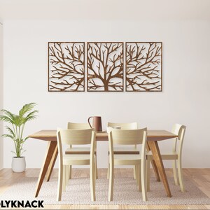 May include: Three-panel wooden wall art featuring a tree branch design. The artwork is a warm brown color and is mounted on a white wall above a wooden dining table with four chairs. A plant in a white pot and a pitcher are also visible.