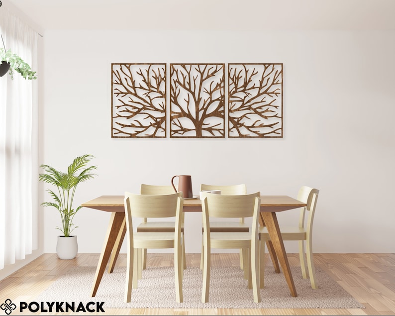 May include: Three-panel wooden wall art featuring a tree branch design. The artwork is a warm brown color and is mounted on a white wall above a wooden dining table with four chairs. A plant in a white pot and a pitcher are also visible.