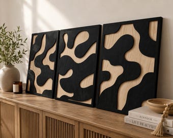 May include: Three abstract wall art panels. Each panel features a black, organic-shaped design against a light wood background. The panels are framed in black and displayed on a wooden cabinet. A vase with greenery, a candle, and books are also visible.
