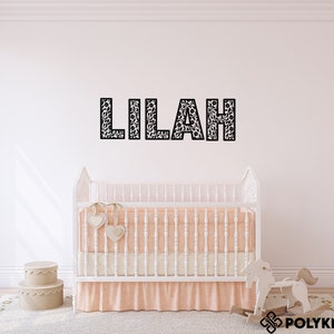 May include: A white crib with a peach-colored skirt and a white rocking horse in front of it. The name "Lilah" is spelled out in black letters on the wall behind the crib.
