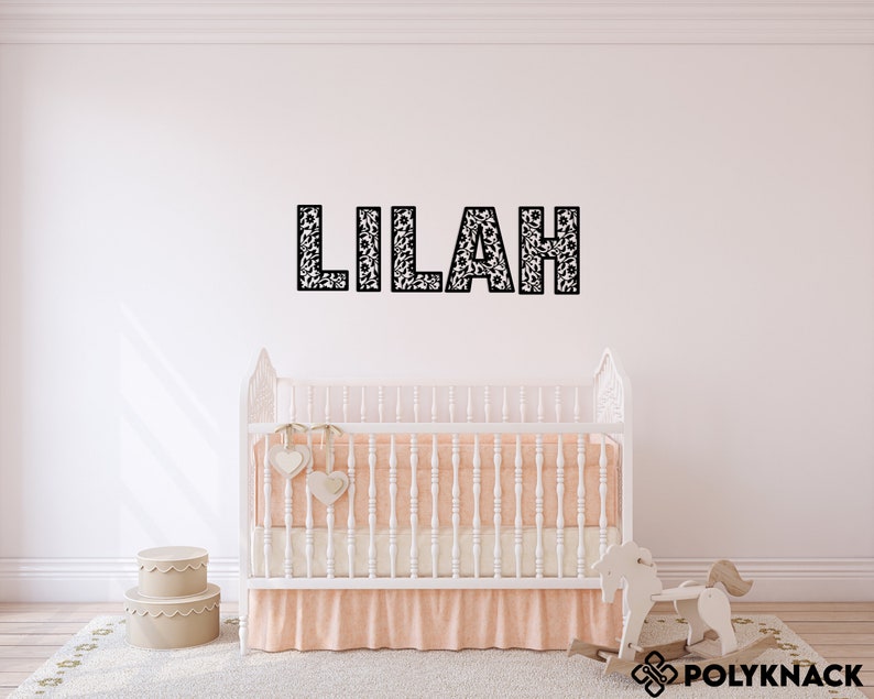 May include: A white crib with a peach-colored skirt and a white rocking horse in front of it. The name "Lilah" is spelled out in black letters on the wall behind the crib.