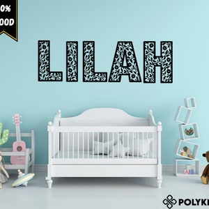 May include: A white crib with a black and white floral patterned name decal that reads "Lilah" on a light blue wall. The decal is made of 100% wood.