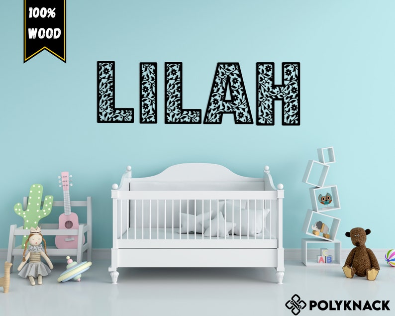 May include: A white crib with a black and white floral patterned name decal that reads "Lilah" on a light blue wall. The decal is made of 100% wood.