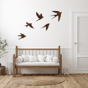 May include: Wooden swallow wall art in various sizes and poses, mounted on a white wall. The birds are a dark brown color, creating a contrast against the white background. A wooden bench with white cushions and a round rug are in the foreground.