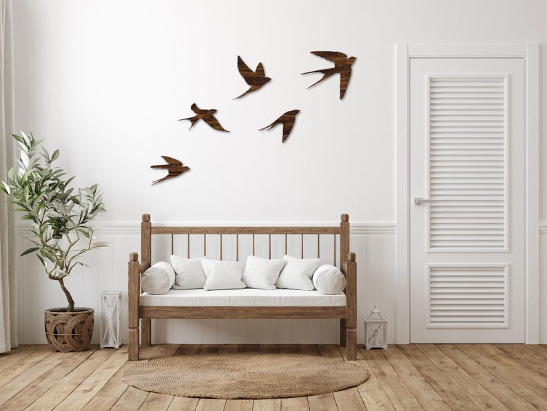 May include: Wooden swallow wall art in various sizes and poses, mounted on a white wall. The birds are a dark brown color, creating a contrast against the white background. A wooden bench with white cushions and a round rug are in the foreground.