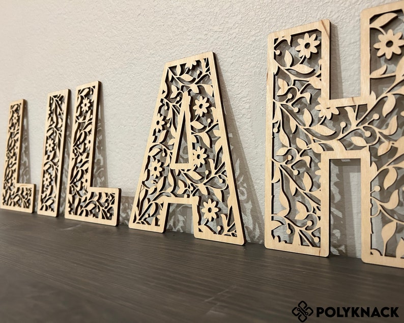 May include: Wooden letters spelling out the name "Lilah" with a floral design cut out. The letters are a light brown color and are arranged in a row on a dark brown surface.