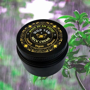 Może przedstawiać: Czarny słoiczek kremu do szyi Wild Yam ze złotymi napisami i motylem. Tekst na wieczku głosi "Made by an Artist", "Wild Yam", "Neck Creme", "Luxury", "Vegan" i "Organic". Tło to rozmyty obraz zielonych liści i deszczu.