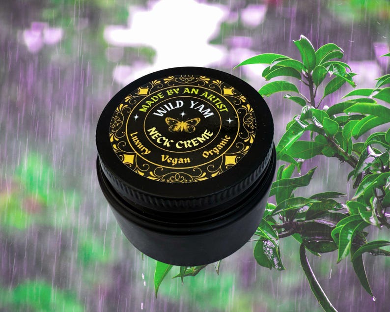 Może przedstawiać: Czarny słoiczek kremu do szyi Wild Yam ze złotymi napisami i motylem. Tekst na wieczku głosi "Made by an Artist", "Wild Yam", "Neck Creme", "Luxury", "Vegan" i "Organic". Tło to rozmyty obraz zielonych liści i deszczu.