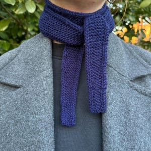 May include: A close-up of a navy blue knitted scarf. The scarf is wrapped around a person's neck and hangs down in two long sections. The scarf is made of a thick, textured yarn and appears to be handmade. The person is wearing a gray coat.