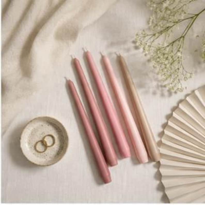 K&ouml;nnte beinhalten: F&uuml;nf Stabkerzen in einem Farbverlauf von Rosa- und Beiget&ouml;nen, die diagonal angeordnet sind. Neben den Kerzen befindet sich eine kleine, runde, gesprenkelte Keramikschale mit zwei goldfarbenen Metallringen. Die Dochte sind oben sichtbar.