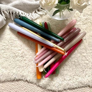 K&ouml;nnte beinhalten: Eine Sammlung langer, schlanker Stabkerzen in verschiedenen Farben, darunter Dunkelgr&uuml;n, Blau, Gold, Orange, Pink, Magenta und Beige. Die Kerzen sind in einem B&uuml;ndel angeordnet und jede verf&uuml;gt &uuml;ber einen kleinen wei&szlig;en Docht am spitzen Ende.