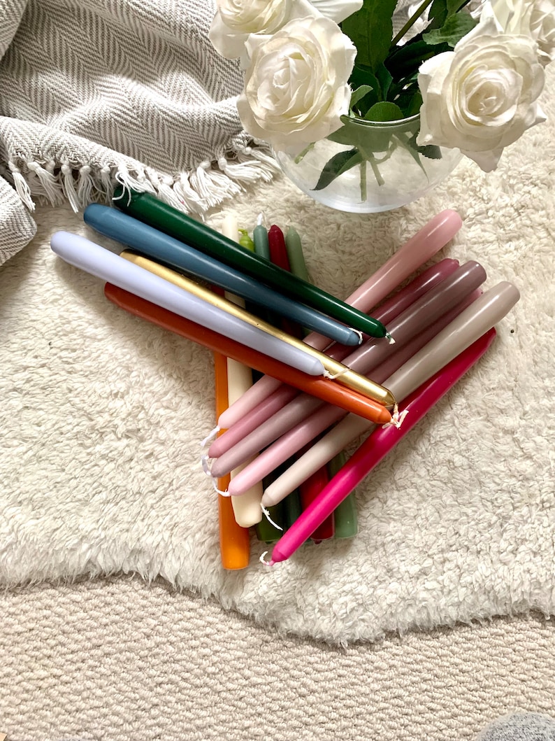 K&ouml;nnte beinhalten: Eine Sammlung langer, schlanker Stabkerzen in verschiedenen Farben, darunter Dunkelgr&uuml;n, Blau, Gold, Orange, Pink, Magenta und Beige. Die Kerzen sind in einem B&uuml;ndel angeordnet und jede verf&uuml;gt &uuml;ber einen kleinen wei&szlig;en Docht am spitzen Ende.