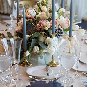 K&ouml;nnte beinhalten: Ein Tischaufsatz mit einem Blumenarrangement aus rosa und wei&szlig;en Bl&uuml;ten, gr&uuml;nem Blattwerk und Sukkulenten, umgeben von hohen schieferblauen Stabkerzen in goldfarbenen Metallhaltern, mit klaren Gl&auml;sern und wei&szlig;en Tellern auf einem Tisch.