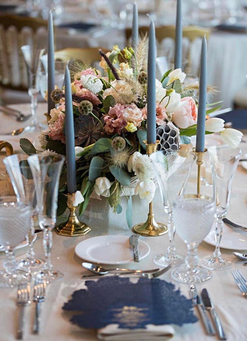 K&ouml;nnte beinhalten: Ein Tischaufsatz mit einem Blumenarrangement aus rosa und wei&szlig;en Bl&uuml;ten, gr&uuml;nem Blattwerk und Sukkulenten, umgeben von hohen schieferblauen Stabkerzen in goldfarbenen Metallhaltern, mit klaren Gl&auml;sern und wei&szlig;en Tellern auf einem Tisch.