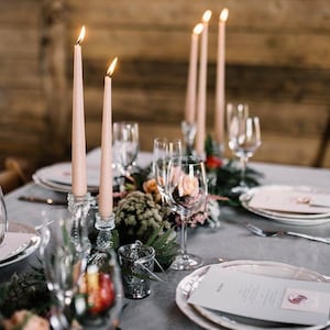 K&ouml;nnte beinhalten: Ein gedeckter Tisch mit hellrosa Stabkerzen in klaren Glashaltern, Weingl&auml;sern, wei&szlig;en Tellern und bedruckten Men&uuml;karten mit Wachssiegel. Das Arrangement umfasst gr&uuml;nes Blattwerk und kleine florale Akzente auf einer grauen Tischdecke.
