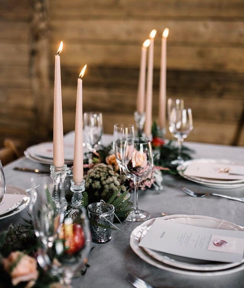 K&ouml;nnte beinhalten: Ein gedeckter Tisch mit hellrosa Stabkerzen in klaren Glashaltern, Weingl&auml;sern, wei&szlig;en Tellern und bedruckten Men&uuml;karten mit Wachssiegel. Das Arrangement umfasst gr&uuml;nes Blattwerk und kleine florale Akzente auf einer grauen Tischdecke.