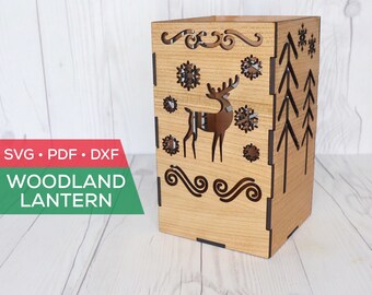May include: A wooden lantern with a cut-out design of a deer, snowflakes, and pine trees. The lantern is made of light brown wood and has a square shape. The text "SVG • PDF • DXF WOODLAND LANTERN" is printed on the bottom of the lantern.