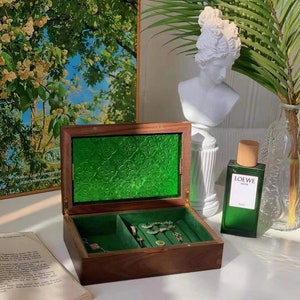 May include: A wooden jewelry box with a green velvet interior and a green glass lid. The box is open and contains jewelry, including a necklace, earrings, and rings. The box is sitting on a white surface with a bottle of perfume and a white bust sculpture.