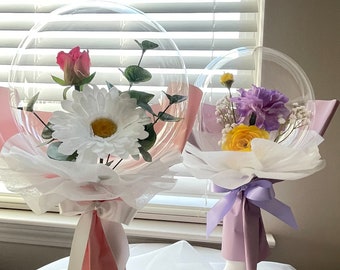 Peut inclure: Deux bouquets de bulles transparentes avec du papier de soie blanc et un ruban. Le bouquet de gauche a un ruban rose et présente une marguerite blanche, une rose rose et des feuilles d'eucalyptus. Le bouquet de droite a un ruban violet et présente une rose jaune, un œillet violet et de la gypsophile blanche.