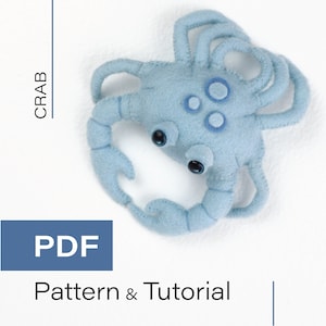 May include: A blue felt crab with black eyes and white circles on its back. The crab is lying on a white surface. PDF Pattern & Tutorial.