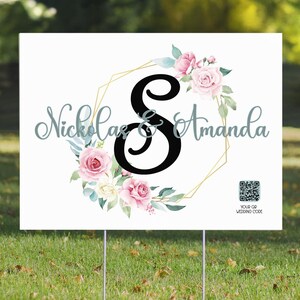 May include: A white wedding sign with a gold geometric frame and floral wreath. The wreath features pink and white roses, green leaves, and a black letter "S" in the centre. The text "Nicholas & Amanda" is written in a cursive font below the wreath. A QR code is in the bottom right corner with the text "Your QR Wedding Code".