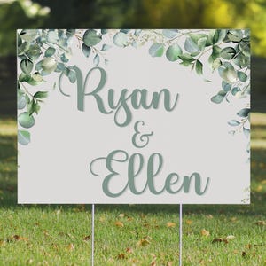 May include: A white wedding sign with green eucalyptus leaves and the text "Ryan & Ellen" in a green script font.