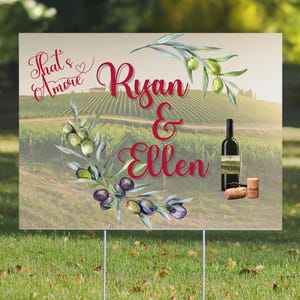 May include: A wedding sign with a vineyard background and olive branches. The sign says "That's Amore" with a heart and "Ryan & Ellen" in red cursive. There is a bottle of wine and two corks on the sign.