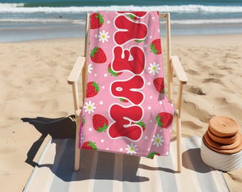 May include: A pink beach towel with the name "MAYA" in large red bubble letters. The towel features a pattern of strawberries and white daisies. The towel is draped over a wooden beach chair on a sandy beach.