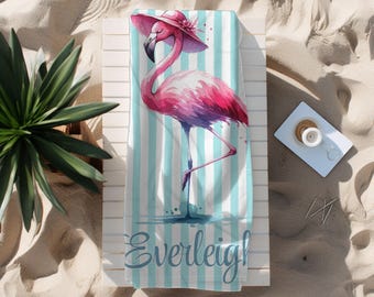 May include: A beach towel with a flamingo illustration wearing a pink hat, set against a blue and white striped background. The flamingo is pink and purple, with the name "Everleigh!" in script at the bottom.