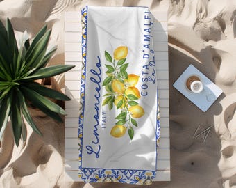 May include: White beach towel with a blue and yellow border, featuring a lemon branch illustration and the words "Limoncello Italy Costa D'Amalfi". The towel is folded on a white wooden surface, with sand in the background.