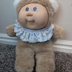 Peut inclure: Une poup&eacute;e Cabbage Patch Kids vintage avec un corps marron en peluche et un visage doux et rond. La poup&eacute;e a les yeux bleus, une expression souriante et un col &agrave; volants avec un motif pastel. La poup&eacute;e porte une capuche marron en forme d'ours avec des accents d'oreilles blanches.