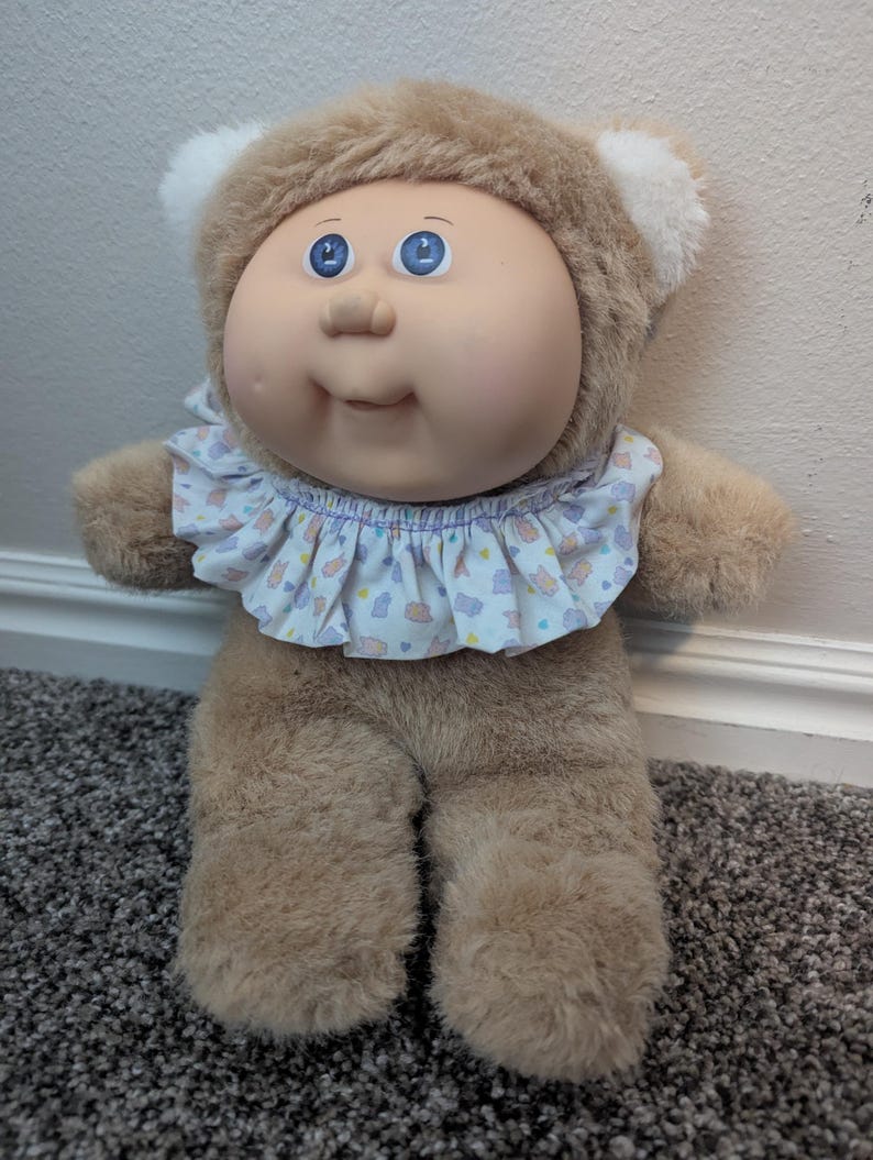 Peut inclure: Une poup&eacute;e Cabbage Patch Kids vintage avec un corps marron en peluche et un visage doux et rond. La poup&eacute;e a les yeux bleus, une expression souriante et un col &agrave; volants avec un motif pastel. La poup&eacute;e porte une capuche marron en forme d'ours avec des accents d'oreilles blanches.