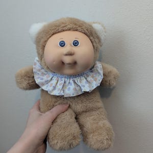 May include: A vintage Cabbage Patch Kids doll with a brown bear-like hood and plush body. The doll has a round face with blue eyes and a ruffled collar with a pastel pattern. The doll is a collectible item.
