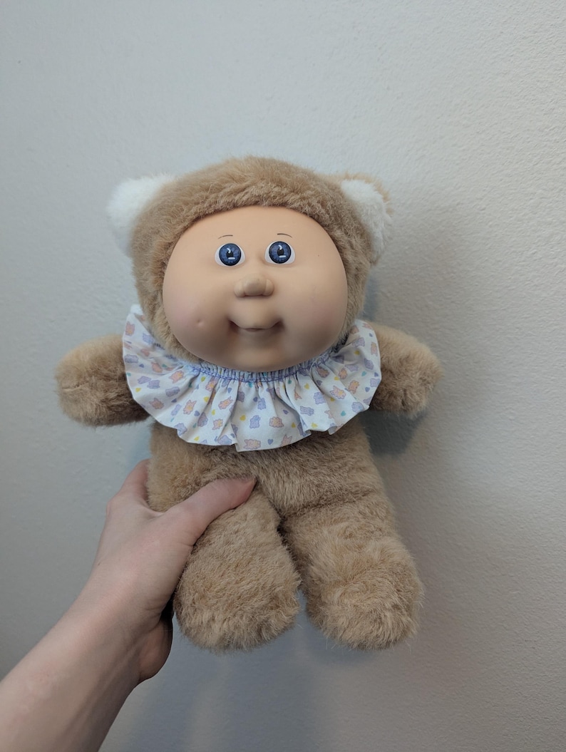 Peut inclure: Poup&eacute;e Cabbage Patch Kids vintage avec une capuche en forme d'ours marron et un corps en peluche. La poup&eacute;e a un visage rond avec des yeux bleus et un col &agrave; volants avec un motif pastel. C'est un objet de collection.