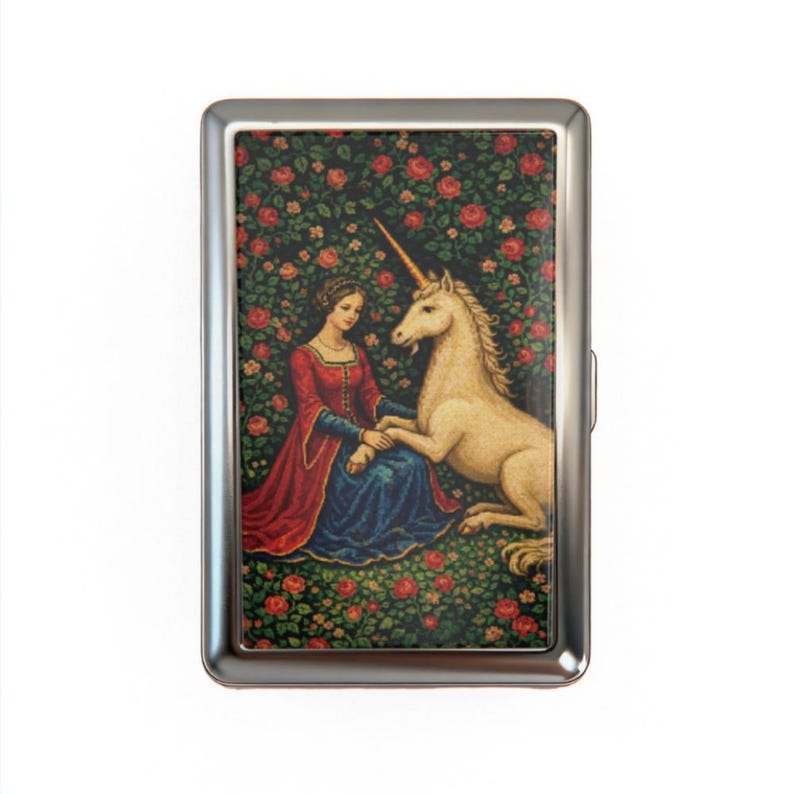 May include: A silver metal cigarette case featuring a medieval scene. The artwork depicts a woman in a red and blue dress seated next to a white unicorn, surrounded by a rose garden. The case has a rectangular shape.