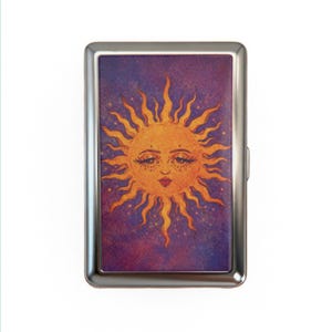 Puede incluir: Un estuche de cigarrillos de metal plateado con una ilustración de sol caprichosa. El sol tiene una cara sonriente con largas pestañas y un labio rojo, sobre un fondo estrellado púrpura y azul. Los rayos del sol son de color naranja brillante.
