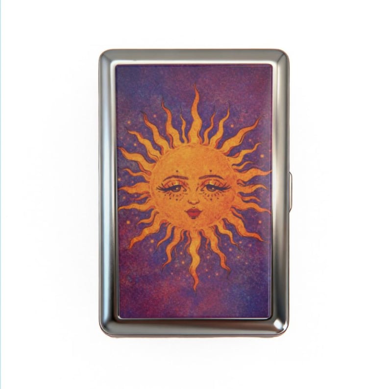May include: A silver metal cigarette case featuring a whimsical sun illustration. The sun has a smiling face with long eyelashes and a red lip, set against a purple and blue starry background. The sun's rays are a bright orange color.