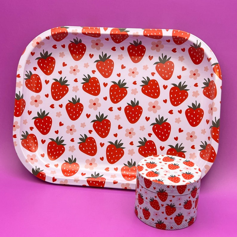 Op de afbeelding: Een witte metalen dienblad met een roze achtergrond en een aardbeienpatroon. Het dienblad is gevuld met rode aardbeien en heeft een witte metalen deksel met een aardbeienpatroon. Het deksel ligt op het dienblad.