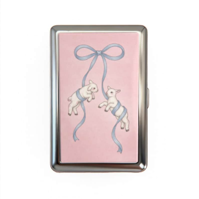 May include: A silver metal cigarette case with a pink background. The case features a watercolor illustration of two white lambs wrapped in blue ribbons. A blue ribbon bow is at the top of the case.