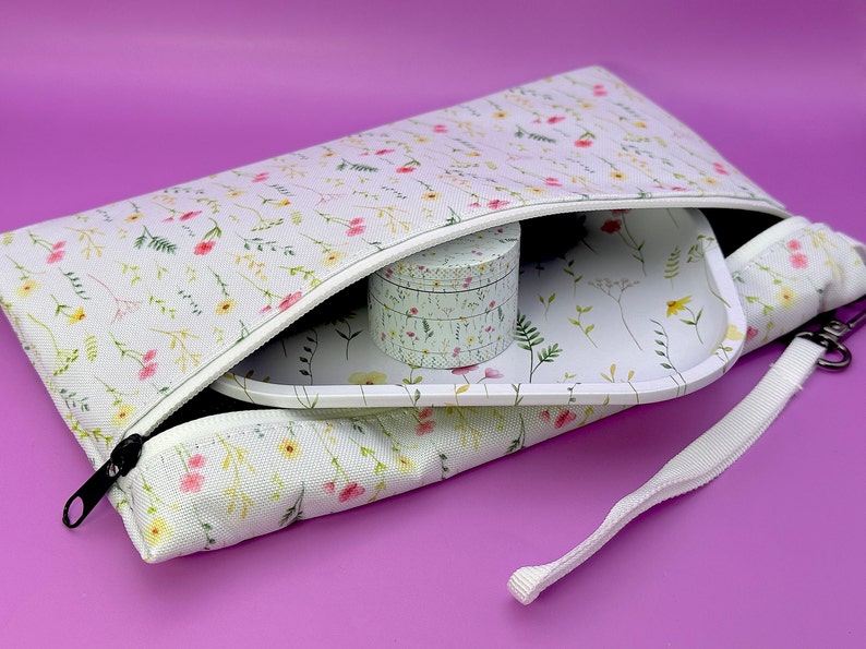 Peut inclure: Une pochette blanche &agrave; fermeture &eacute;clair avec un imprim&eacute; floral et une sangle blanche. La pochette est ouverte et contient un plateau en c&eacute;ramique blanche et un r&eacute;cipient rond blanc avec un motif floral.