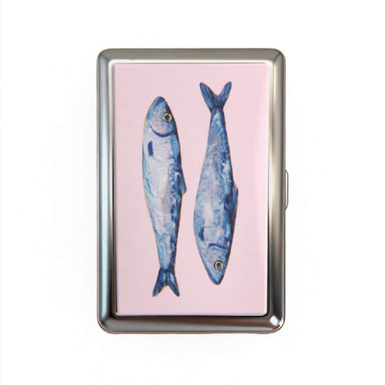 May include: A silver metal cigarette case with a pink background. The case features a watercolor painting of two blue fish. The fish are oriented vertically, with one facing up and the other facing down.