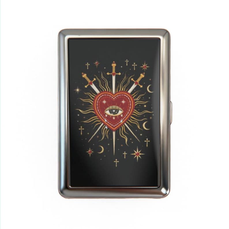 May include: A silver metal cigarette case with a black background featuring a heart pierced by three swords. The heart has an eye in the center and is surrounded by sun rays, stars, and crescent moons.